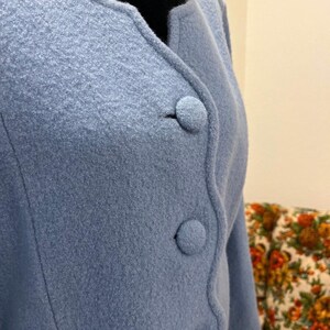 May include: A light blue jacket with three round buttons. The jacket has a rounded collar and a textured fabric. The sleeves are three-quarter length. The background includes a floral patterned chair.