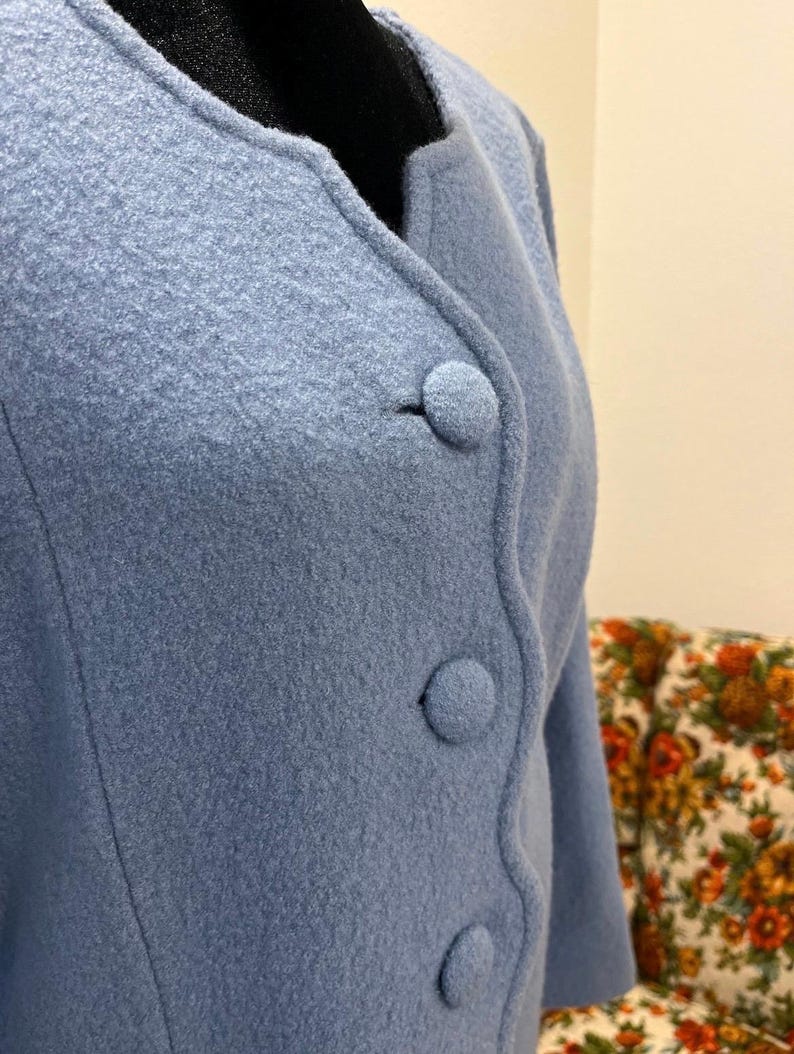 May include: A light blue jacket with three round buttons. The jacket has a rounded collar and a textured fabric. The sleeves are three-quarter length. The background includes a floral patterned chair.