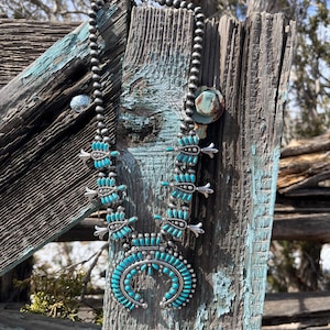 May include: A turquoise and silver Native American style necklace with a crescent moon pendant. The necklace is made of turquoise beads and silver beads and is hanging on a weathered wooden fence post.