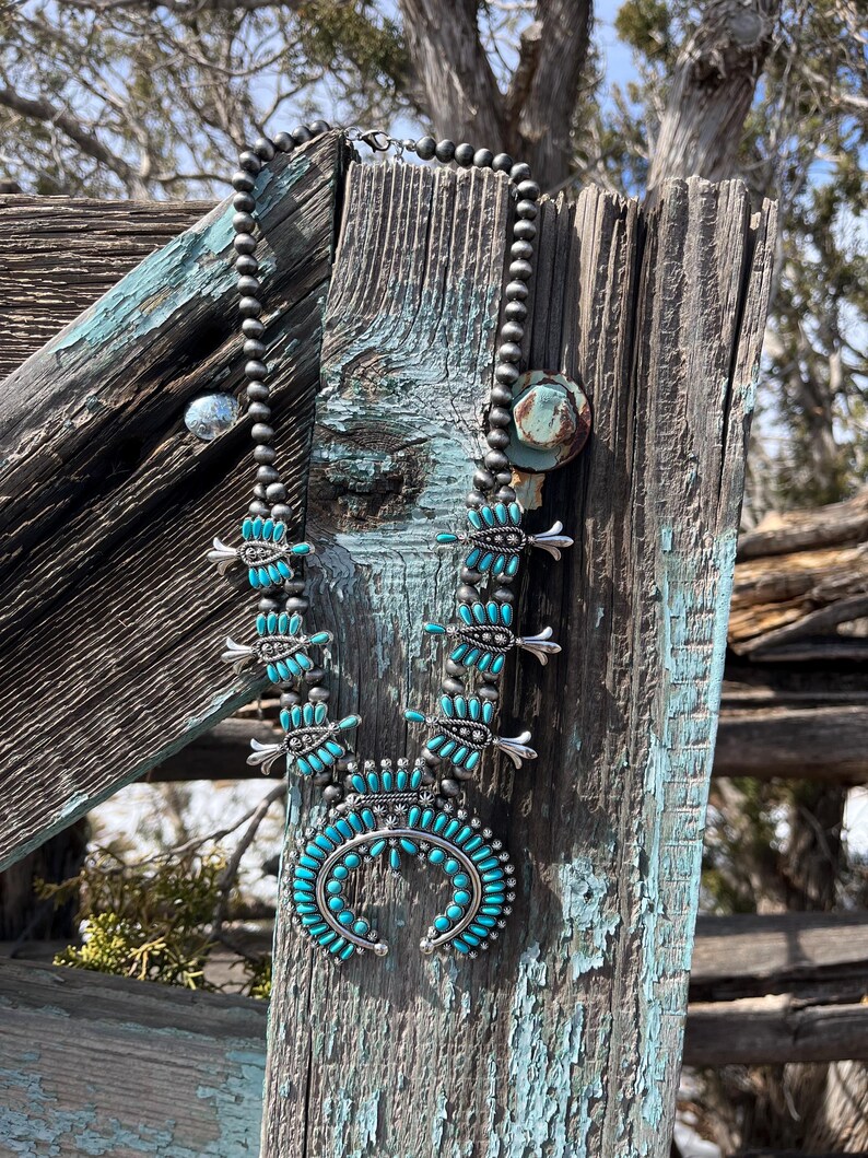 May include: A turquoise and silver Native American style necklace with a crescent moon pendant. The necklace is made of turquoise beads and silver beads and is hanging on a weathered wooden fence post.
