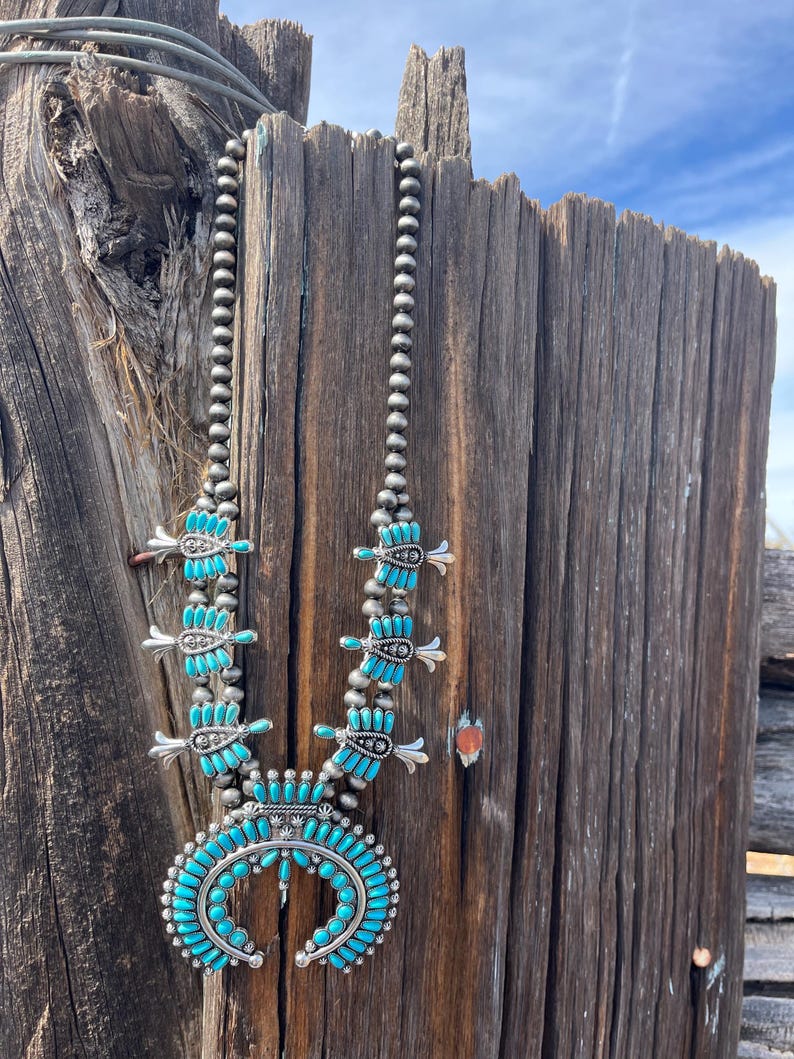 May include: A turquoise and silver Native American necklace with a crescent moon pendant. The necklace is made of turquoise beads and silver beads, and the pendant is a crescent moon shape with a turquoise stone in the center.