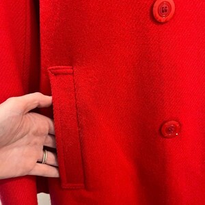 May include: A close-up of a vibrant red coat. The coat features a tailored pocket and two matching buttons. The fabric has a textured appearance. The coat is a solid color, with no visible patterns or designs.