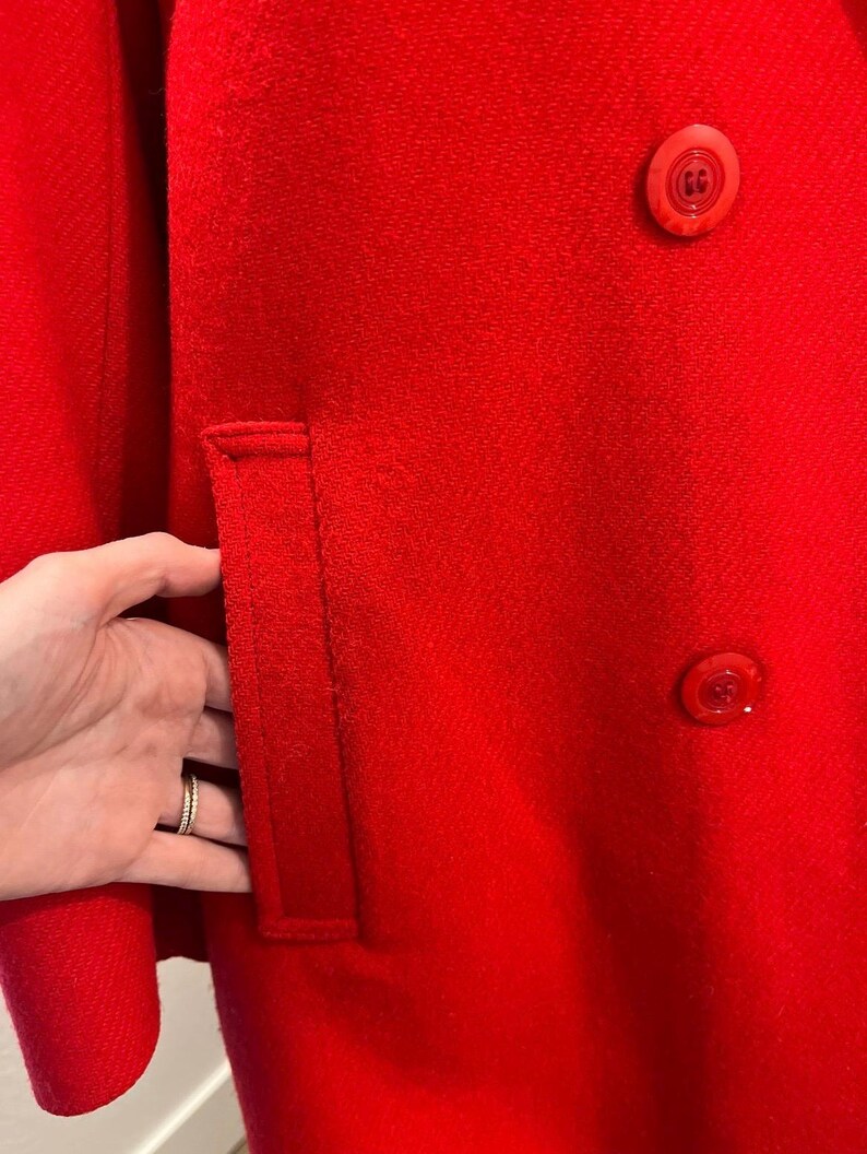 May include: A close-up of a vibrant red coat. The coat features a tailored pocket and two matching buttons. The fabric has a textured appearance. The coat is a solid color, with no visible patterns or designs.