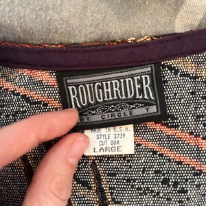 May include: A close-up of a woven fabric with a label that reads "ROUGHRIDER BY CIRCLE T." The label also indicates "MADE IN U.S.A." and "LARGE." The fabric has a pattern of black, orange, and tan.