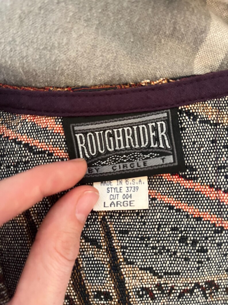 May include: A close-up of a woven fabric with a label that reads "ROUGHRIDER BY CIRCLE T." The label also indicates "MADE IN U.S.A." and "LARGE." The fabric has a pattern of black, orange, and tan.
