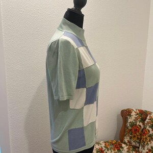 K&ouml;nnte beinhalten: Ein kurz&auml;rmliges Oberteil mit Stehkragen in Hellgr&uuml;n, mit einem Patchwork-Design aus hellblauen, wei&szlig;en und gr&uuml;nen Quadraten. Das Oberteil wird auf einer schwarzen Schaufensterpuppe pr&auml;sentiert.