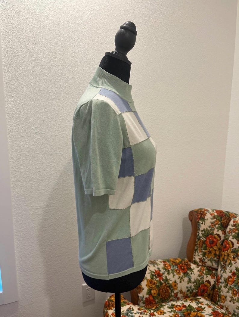K&ouml;nnte beinhalten: Ein kurz&auml;rmliges Oberteil mit Stehkragen in Hellgr&uuml;n, mit einem Patchwork-Design aus hellblauen, wei&szlig;en und gr&uuml;nen Quadraten. Das Oberteil wird auf einer schwarzen Schaufensterpuppe pr&auml;sentiert.