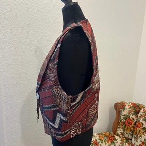 May include: A tapestry vest in shades of red, brown, and black, displayed on a black mannequin. The vest features an intricate woven pattern with geometric and floral motifs. It has a button closure and a decorative fringe.