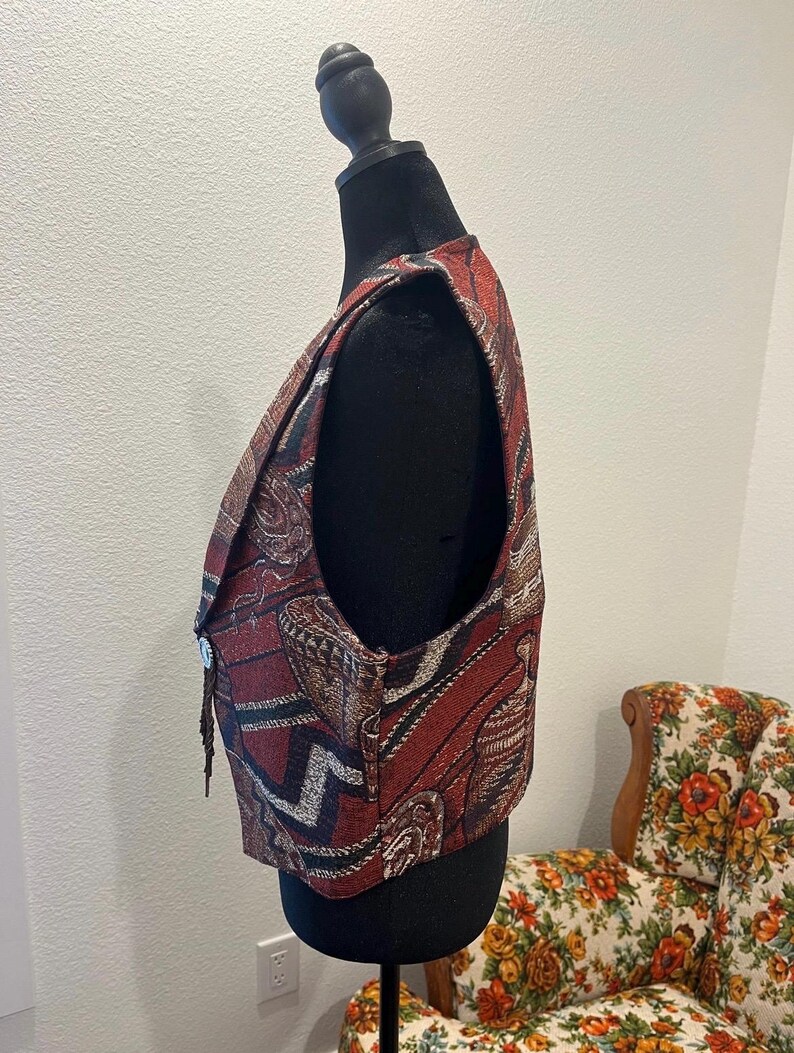 May include: A tapestry vest in shades of red, brown, and black, displayed on a black mannequin. The vest features an intricate woven pattern with geometric and floral motifs. It has a button closure and a decorative fringe.