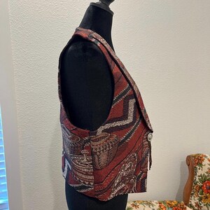 May include: A tapestry vest with a Southwestern-inspired pattern in shades of red, brown, black, and cream. The vest has a button closure and a decorative tassel. The vest is displayed on a black mannequin.