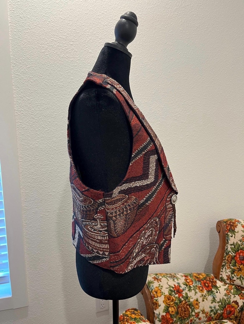 May include: A tapestry vest with a Southwestern-inspired pattern in shades of red, brown, black, and cream. The vest has a button closure and a decorative tassel. The vest is displayed on a black mannequin.