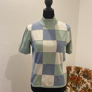 K&ouml;nnte beinhalten: Ein kurz&auml;rmliger Rollkragenpullover im Patchwork-Design. Der Pullover hat Quadrate in Hellblau, Off-White und Hellgr&uuml;n. Das Kleidungsst&uuml;ck wird auf einer schwarzen Schaufensterpuppe pr&auml;sentiert.