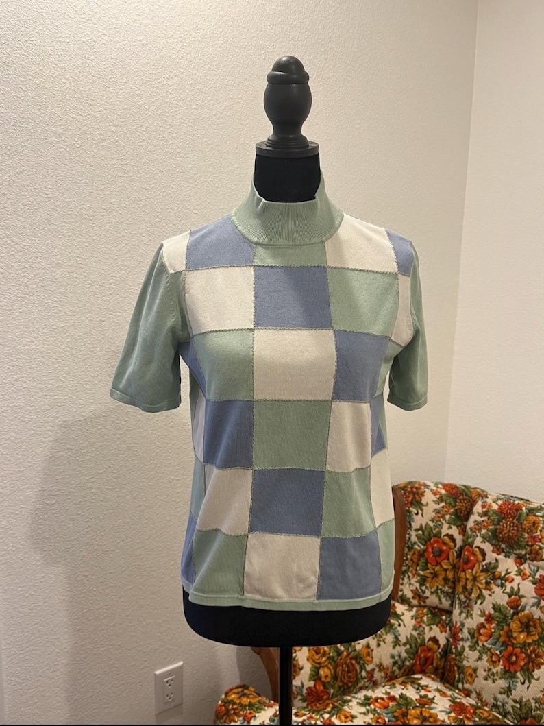 K&ouml;nnte beinhalten: Ein kurz&auml;rmliger Rollkragenpullover im Patchwork-Design. Der Pullover hat Quadrate in Hellblau, Off-White und Hellgr&uuml;n. Das Kleidungsst&uuml;ck wird auf einer schwarzen Schaufensterpuppe pr&auml;sentiert.