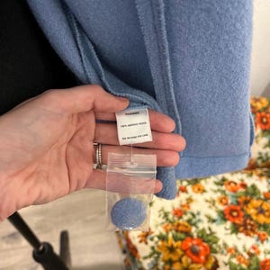 May include: A light blue wool coat with a tag that reads "100% Merino Wool." A small sample of the coat's material is included in a clear plastic bag. The coat is hanging on a black stand.