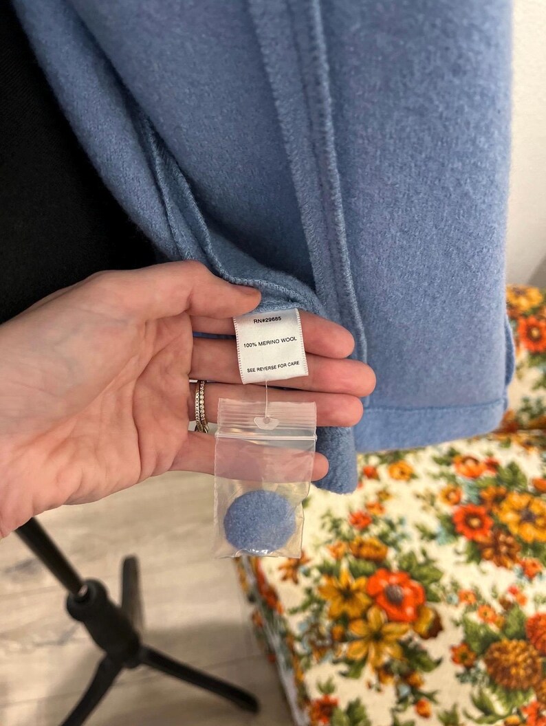 May include: A light blue wool coat with a tag that reads "100% Merino Wool." A small sample of the coat's material is included in a clear plastic bag. The coat is hanging on a black stand.