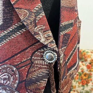 May include: A tapestry vest with a red, brown, and gold pattern. The vest has silver-coloured buttons and brown braided leather accents. The vest is displayed on a black mannequin.
