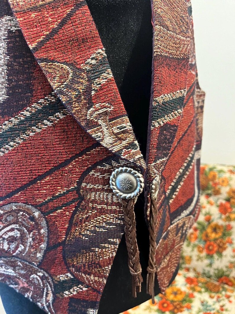 May include: A tapestry vest with a red, brown, and gold pattern. The vest has silver-coloured buttons and brown braided leather accents. The vest is displayed on a black mannequin.