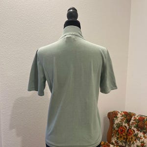 K&ouml;nnte beinhalten: Hellgr&uuml;nes Kurzarm-Shirt mit Stehkragen, pr&auml;sentiert auf einer schwarzen Schaufensterpuppe. Das Shirt hat ein schlichtes, klassisches Design und ist aus einem weichen Strickstoff gefertigt. Der Hintergrund umfasst einen floral gemusterten Stuhl.