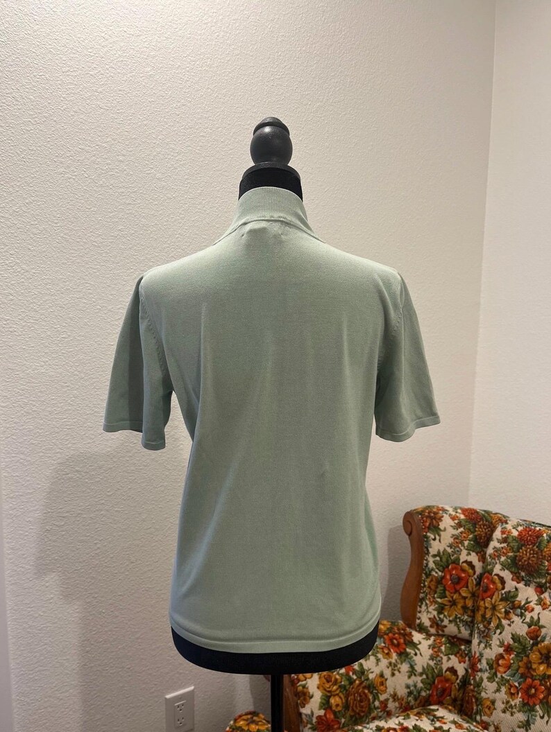 K&ouml;nnte beinhalten: Hellgr&uuml;nes Kurzarm-Shirt mit Stehkragen, pr&auml;sentiert auf einer schwarzen Schaufensterpuppe. Das Shirt hat ein schlichtes, klassisches Design und ist aus einem weichen Strickstoff gefertigt. Der Hintergrund umfasst einen floral gemusterten Stuhl.