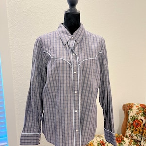 May include: A long-sleeved button-down shirt featuring a blue and white plaid pattern. The shirt includes white piping along the front placket, collar, and western-style shoulder yokes. It has a standard collar and cuffs with small white buttons.