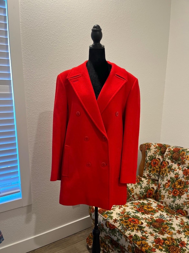 May include: A vibrant red double-breasted coat displayed on a black mannequin. The coat features a classic notched collar, two rows of buttons, and a tailored fit. The coat is a solid, bright red color.