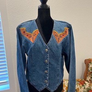 May include: A blue denim jacket with a V-neck and long sleeves. The jacket features decorative orange and yellow embroidered accents on the shoulders and a button-up front. The jacket is displayed on a black mannequin.