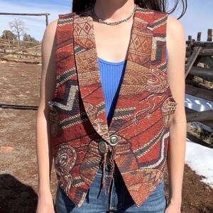 May include: A tapestry vest with a Southwestern pattern in shades of red, brown, and black. The vest has a notched collar, a decorative silver button closure, and a brown leather bolo tie. The person is wearing a blue vest top and blue jeans.