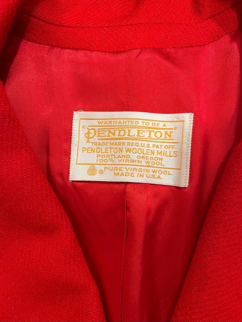 May include: Close-up of a bright red jacket interior. The lining is a matching red. A white label with gold lettering reads "Warranted to be a Pendleton" and details about the wool and origin, "Made in U.S.A."