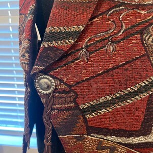 May include: Close-up of a tapestry vest in shades of red, brown, and black. The vest features a detailed woven pattern with silver-toned buttons and braided leather accents. The design has a Southwestern or Western theme.