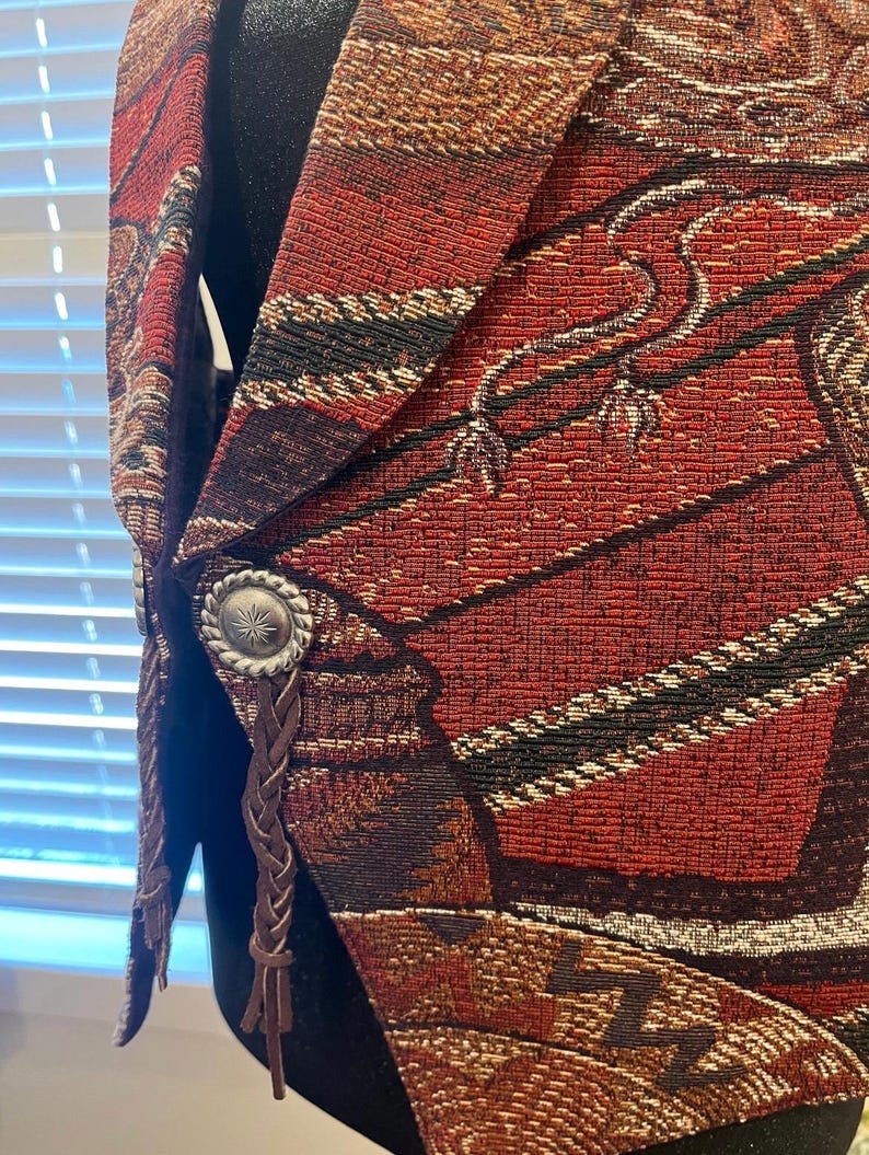 May include: Close-up of a tapestry vest in shades of red, brown, and black. The vest features a detailed woven pattern with silver-toned buttons and braided leather accents. The design has a Southwestern or Western theme.