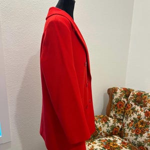 May include: A vibrant red blazer displayed on a black mannequin. The double-breasted jacket features a classic lapel and buttons. The blazer is a solid color, and the fabric appears to be a medium weight. The background includes a floral patterned chair.