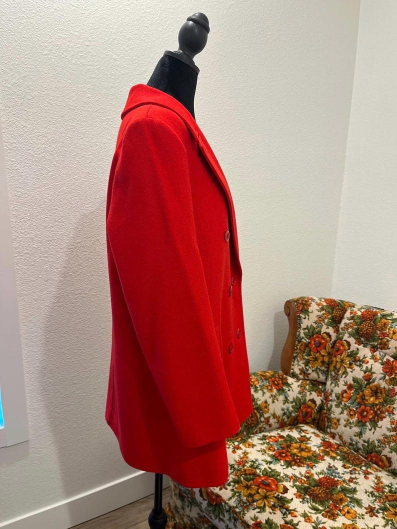 May include: A vibrant red blazer displayed on a black mannequin. The double-breasted jacket features a classic lapel and buttons. The blazer is a solid color, and the fabric appears to be a medium weight. The background includes a floral patterned chair.