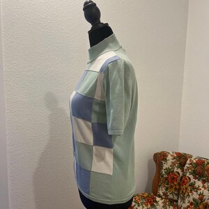 K&ouml;nnte beinhalten: Ein hellgr&uuml;ner Kurzarmpullover mit Stehkragen. Die Vorderseite des Pullovers zeigt ein Patchwork-Design aus hellblauen, wei&szlig;en und gr&uuml;nen Quadraten. Der Pullover wird auf einer schwarzen Schaufensterpuppe pr&auml;sentiert.