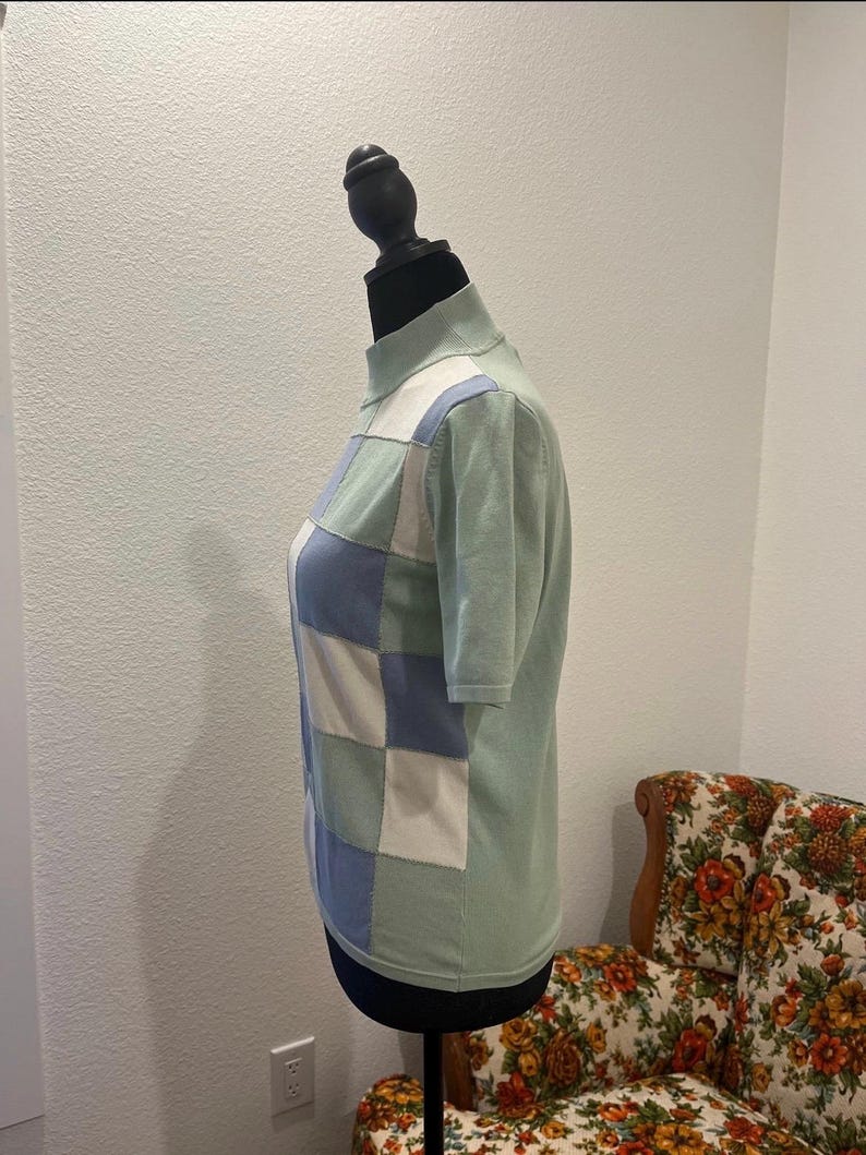 K&ouml;nnte beinhalten: Ein hellgr&uuml;ner Kurzarmpullover mit Stehkragen. Die Vorderseite des Pullovers zeigt ein Patchwork-Design aus hellblauen, wei&szlig;en und gr&uuml;nen Quadraten. Der Pullover wird auf einer schwarzen Schaufensterpuppe pr&auml;sentiert.