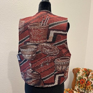 May include: A tapestry vest with a repeating pattern of Southwestern-style baskets and pottery in red, brown, and cream. The vest is displayed on a black mannequin, highlighting its unique design and texture. The vest has a V-neck and a tailored fit.