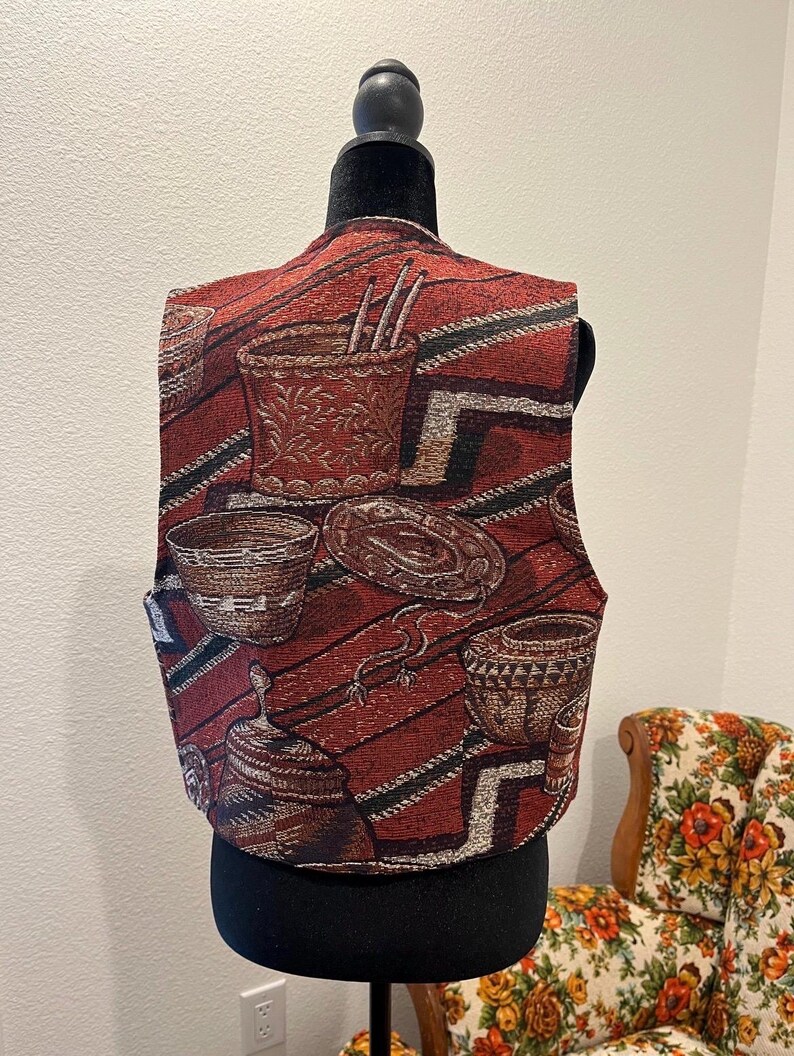 May include: A tapestry vest with a repeating pattern of Southwestern-style baskets and pottery in red, brown, and cream. The vest is displayed on a black mannequin, highlighting its unique design and texture. The vest has a V-neck and a tailored fit.