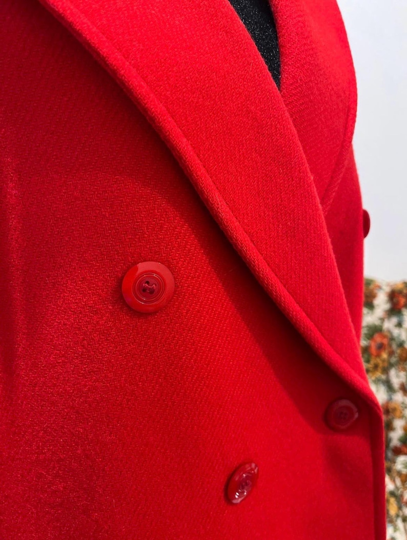 May include: Close-up of a bright red double-breasted blazer. The blazer features matching red buttons and a tailored lapel. The fabric has a subtle texture, and the overall design is classic and stylish.