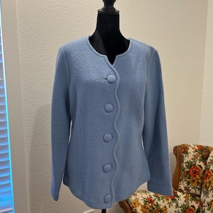 May include: A light blue jacket with a rounded neckline and five large, matching buttons down the front. The jacket is displayed on a black mannequin. A floral patterned armchair is visible in the background.
