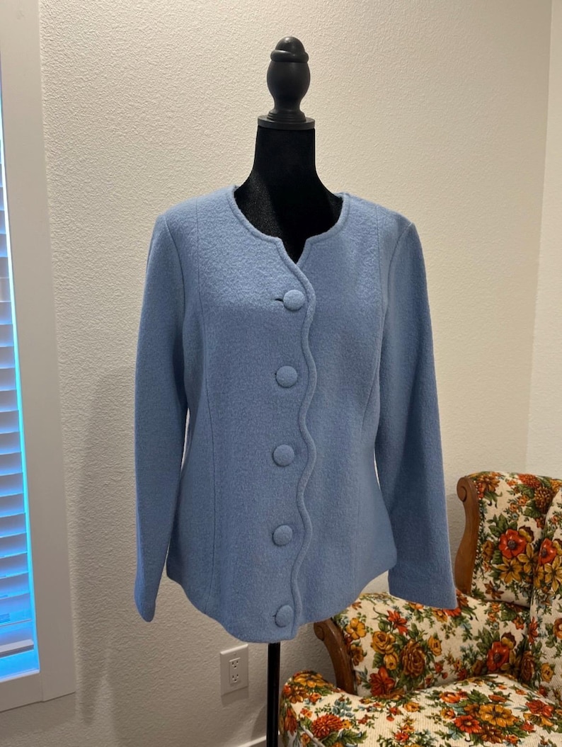May include: A light blue jacket with a rounded neckline and five large, matching buttons down the front. The jacket is displayed on a black mannequin. A floral patterned armchair is visible in the background.