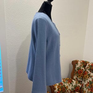 May include: A light blue jacket with long sleeves and a round neckline. The jacket is displayed on a black mannequin. The fabric appears to be a soft, textured material. The jacket has a button closure.