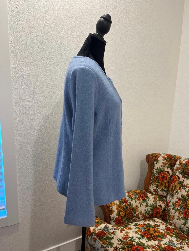 May include: A light blue jacket with long sleeves and a round neckline. The jacket is displayed on a black mannequin. The fabric appears to be a soft, textured material. The jacket has a button closure.