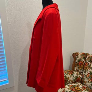 May include: A vibrant red coat displayed on a black mannequin. The coat features a classic collar, two buttons, and a tailored silhouette. The coat is made of a soft, possibly wool, material. The coat is a medium length.
