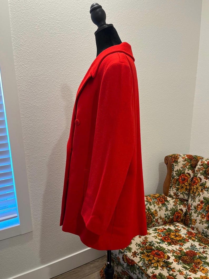 May include: A vibrant red coat displayed on a black mannequin. The coat features a classic collar, two buttons, and a tailored silhouette. The coat is made of a soft, possibly wool, material. The coat is a medium length.