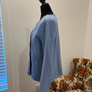 May include: A light blue jacket with a round neckline and button closure. The jacket is displayed on a black mannequin. The background includes a floral patterned armchair and a white wall.