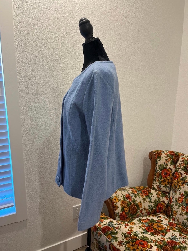 May include: A light blue jacket with a round neckline and button closure. The jacket is displayed on a black mannequin. The background includes a floral patterned armchair and a white wall.