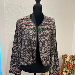 May include: A charcoal grey open-front jacket featuring a repeating cream-coloured geometric pattern. The garment includes horizontal bands of embroidery across the shoulders and cuffs in shades of teal, orange, and white, with a simple round neckline.