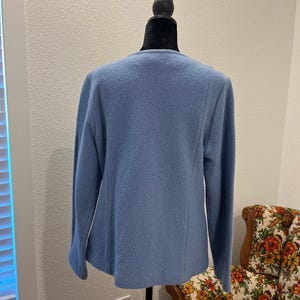 May include: A light blue jacket with long sleeves displayed on a black mannequin. The jacket has a simple, classic design with a round neckline and a slightly textured fabric. The background includes a patterned armchair and a window.
