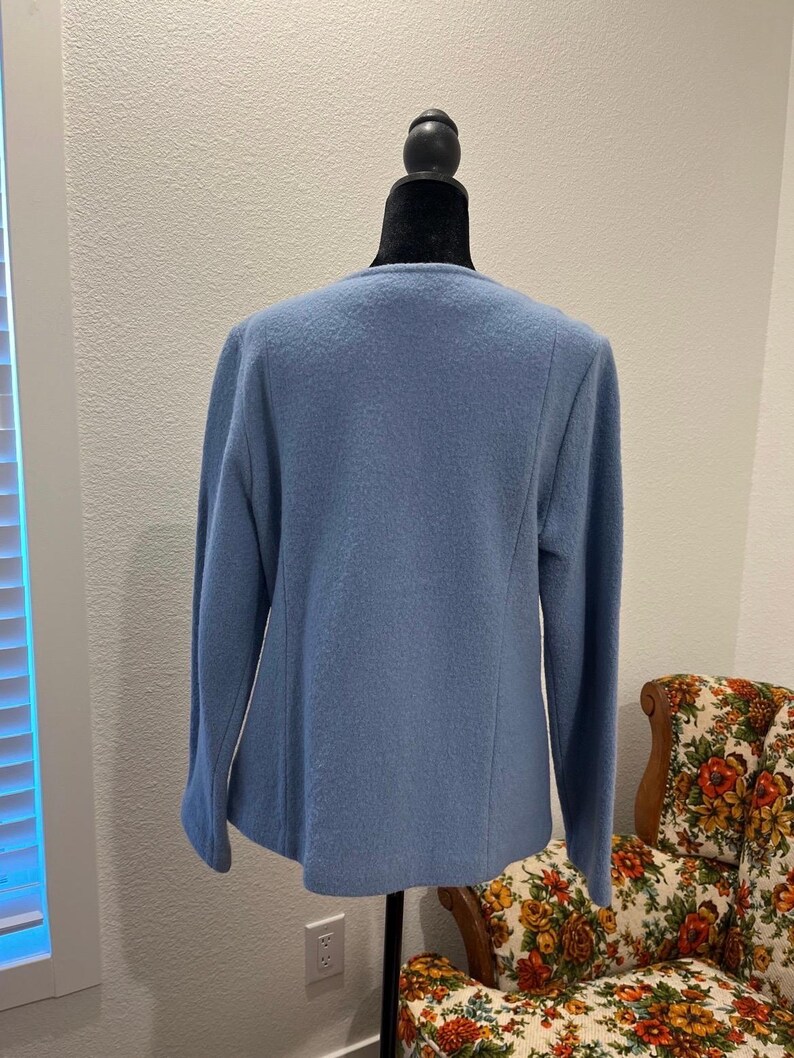 May include: A light blue jacket with long sleeves displayed on a black mannequin. The jacket has a simple, classic design with a round neckline and a slightly textured fabric. The background includes a patterned armchair and a window.