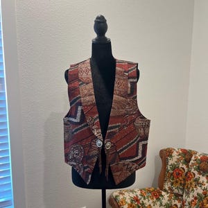May include: A patterned vest in shades of red, brown, and beige, displayed on a black mannequin. The vest features a geometric and tribal design, with two silver-coloured buttons. The vest is a vintage style.