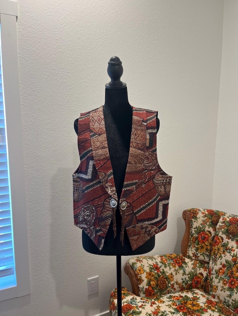 May include: A patterned vest in shades of red, brown, and beige, displayed on a black mannequin. The vest features a geometric and tribal design, with two silver-coloured buttons. The vest is a vintage style.